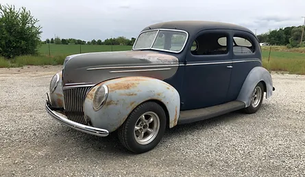 A vintage blue and rusted car parked on a gravel road with a grassy field and overcast sky in the background.<br />
