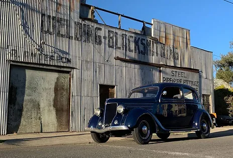 A vintage car parked in front of a rustic industrial building with the sign "GIBB'S BLACKSMITH" and "STEEL PIPE & PIPE FITTINGS"