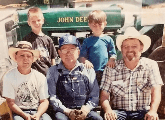 A group of individuals sitting and standing in front of a John Deere tractor, with two of them wearing denim overalls and caps, and one wearing a short-sleeved shirt. The atmosphere suggests a sunny day on a farm.<br />
