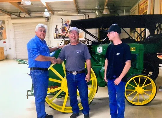 Three men standing in a workshop, two shaking hands, with a classic green and yellow John Deere wagon in the background.