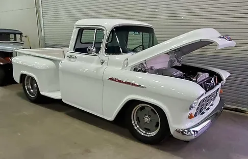 A classic white pickup truck with the hood open, showing a shiny purple engine, parked indoors on a concrete floor.